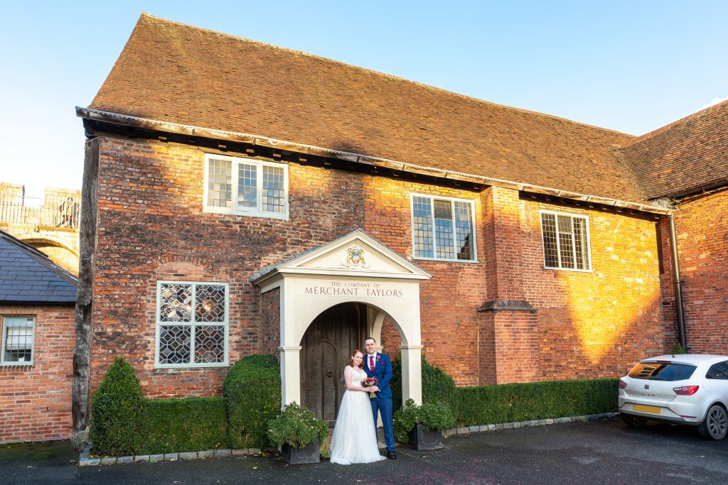 Julia Healey Photography A bride and groom stand smiling under the entrance of a historic brick building labeled "The Courtyard at Merchant Taylors." The scene is sunny, with a white car parked on the right.