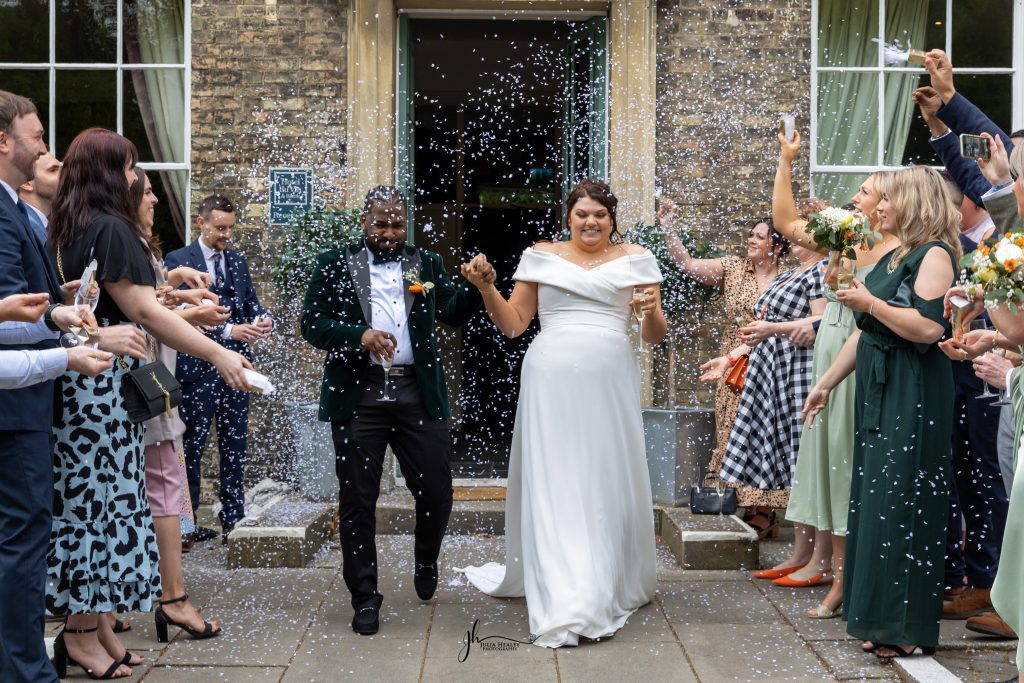 Julia Healey Photography A newlywed couple walks outside as guests throw confetti; the bride wears a white dress and the groom a dark suit, surrounded by people celebrating.