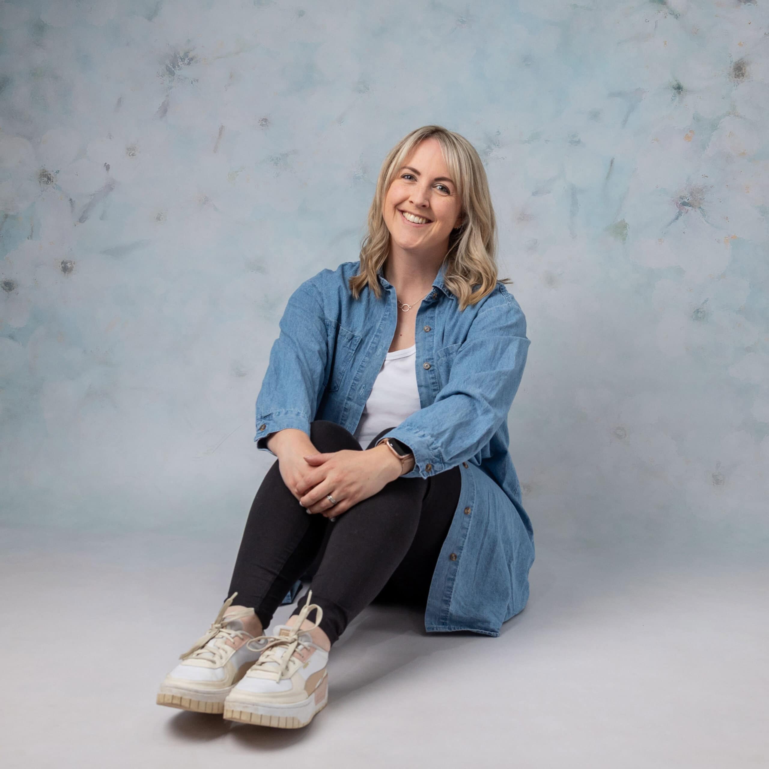 Smiling woman in casual denim outfit, seated on floor.