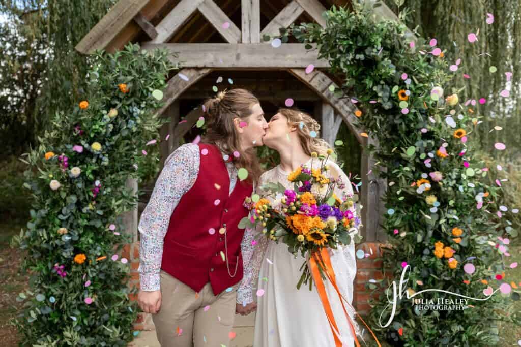 Couple kissing under floral arch with confetti celebration.