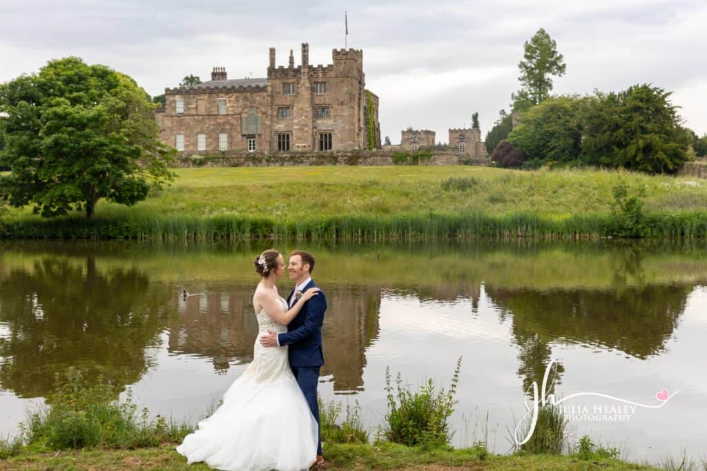 Couple embracing by a serene lake, historic building backdrop.